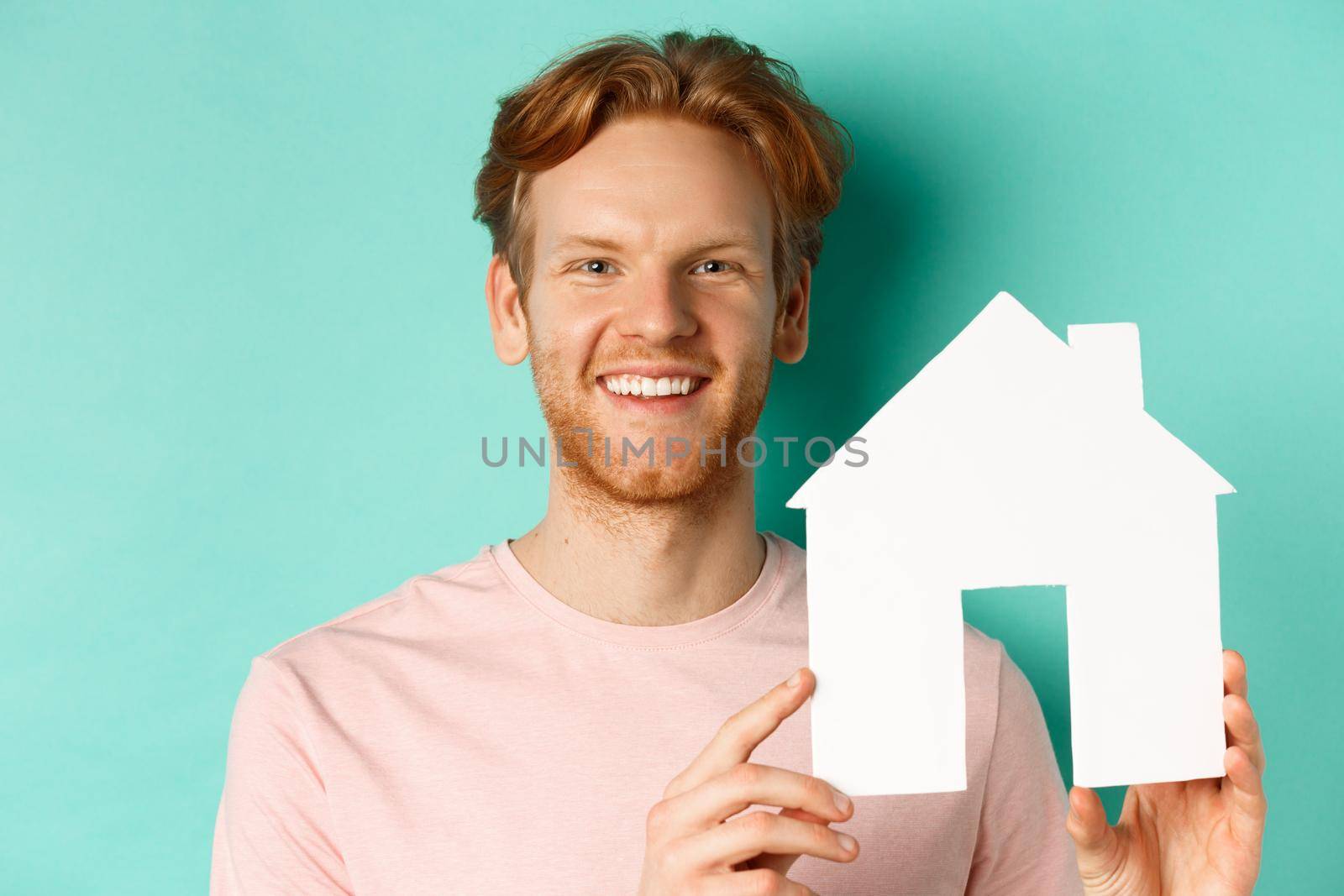 Real estate concept. Close up of redhead bearded man in t-shirt showing paper house cutout, smiling happy at camera, standing over turquoise background.