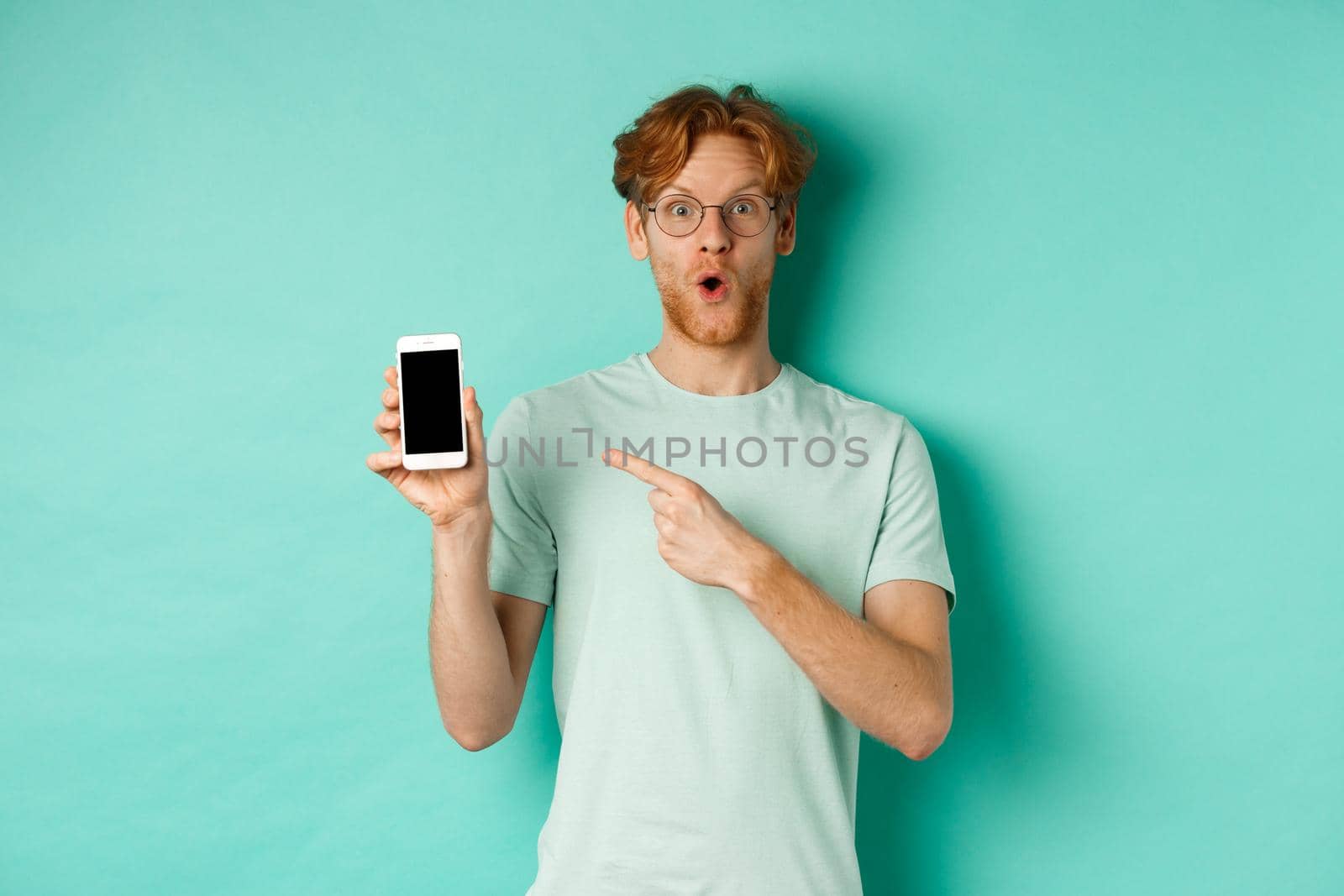Check this out. Handsome redhead guy in glasses pointing finger at blank smartphone screen, showing online promotion, standing amazed over turquoise background by Benzoix