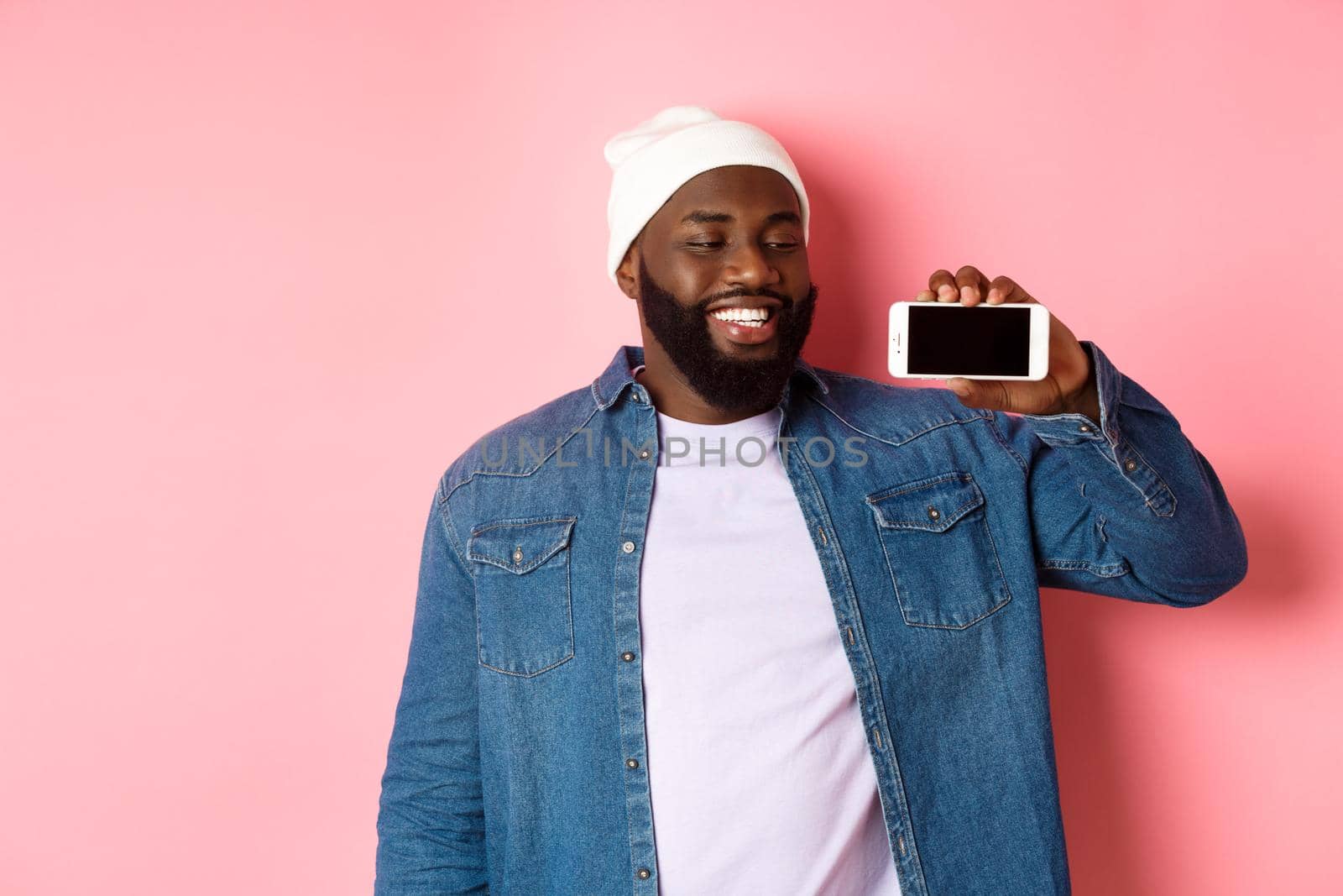 Online shopping and technology concept. Happy young african-american man smiling, showing smartphone screen horizontally with satisfied face expression, pink background.