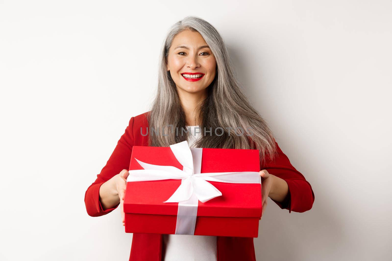 Elegant senior woman giving you present. Asian lady holding red gift box and smiling, standing over white background.