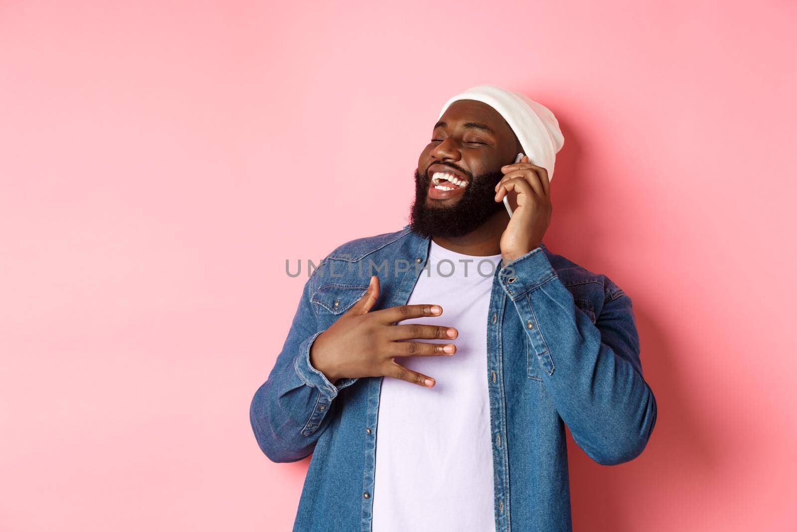 Happy african-american man talking on mobile phone, laughing and smiling, standing in beanie and denim shirt over pink background by Benzoix