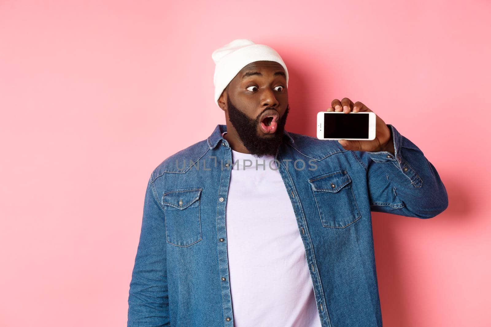 Online shopping and technology concept. Impressed african-american man in beanie showing phone screen, staring at mobile amazed, standing over pink background by Benzoix