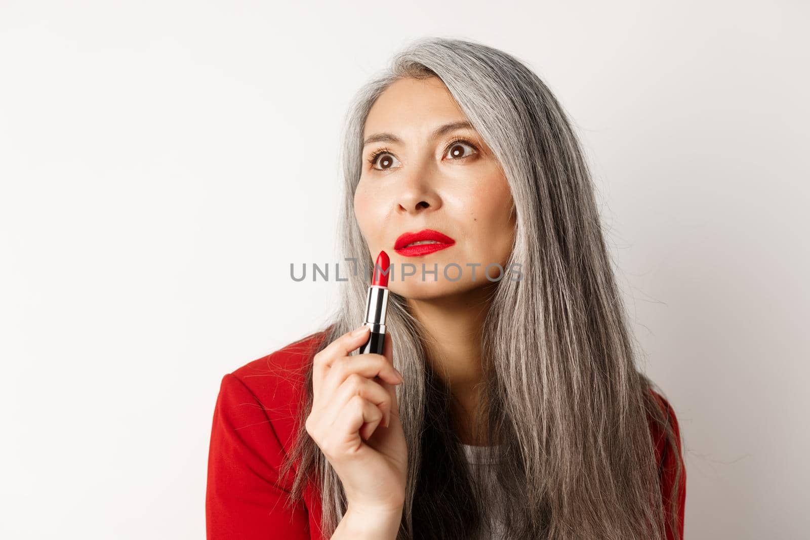 Beauty and makeup concept. Sensual mature asian female with grey hair, looking aside and showing red lipstick, standing over white background by Benzoix