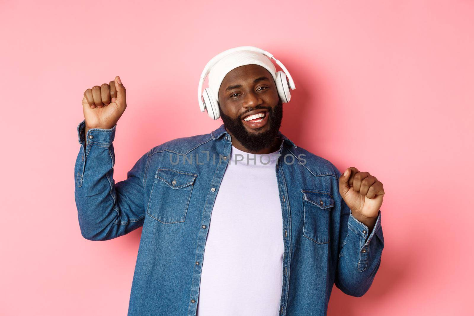 Happy Black man dancing and listening music in headphones, smiling cheerful, standing in streetstyle clothes over pink background.
