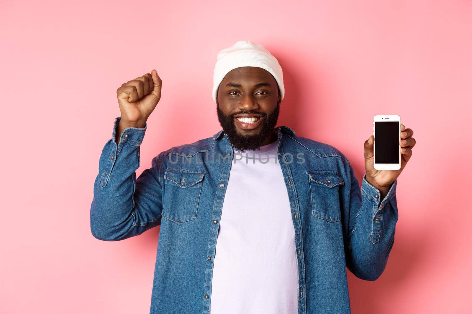 Online shopping and technology concept. Cheerful Black man rejoicing and showing mobile screen, raising hand up satisfied, triumphing while standing over pink background by Benzoix