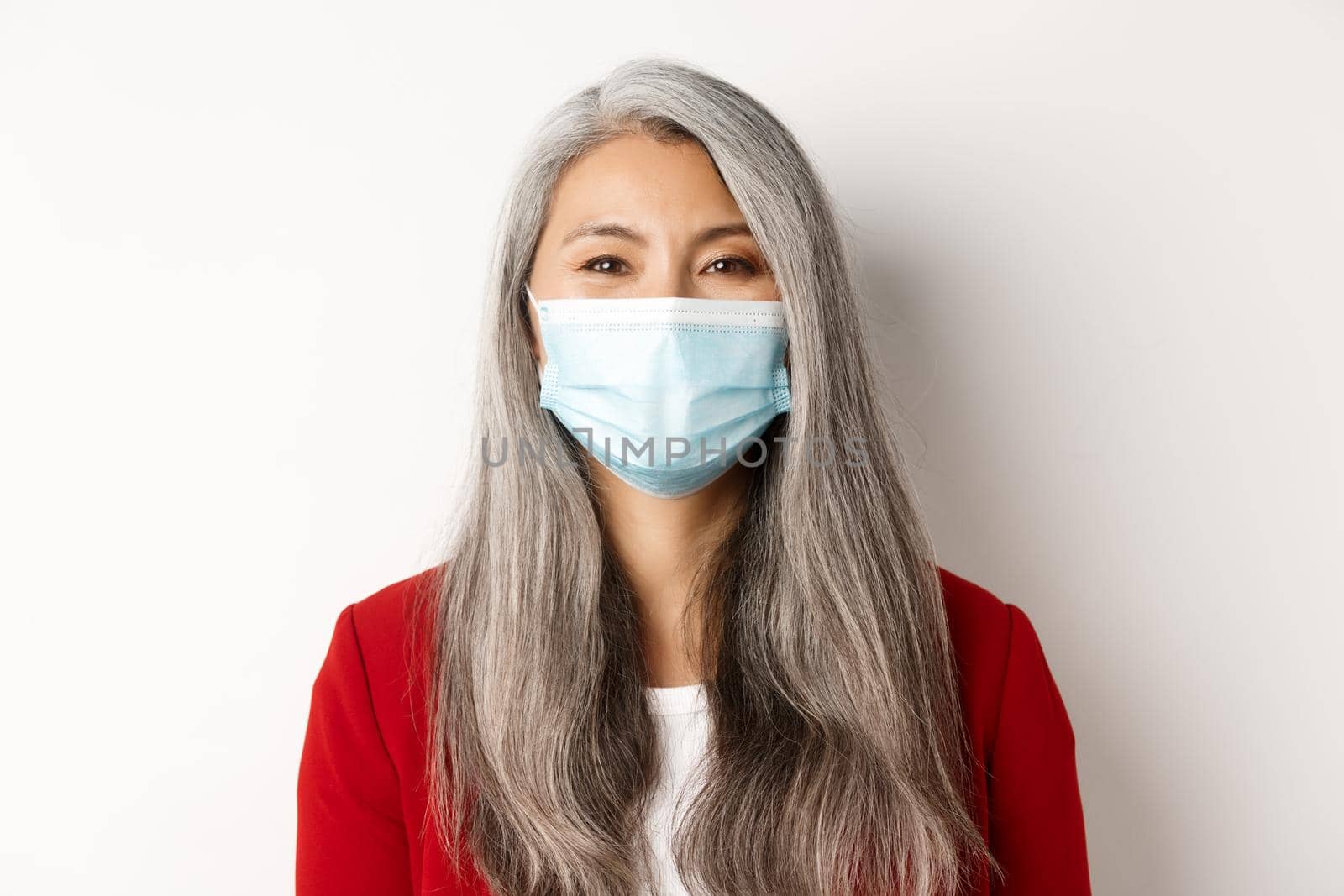 Covid, pandemic and business concept. Close-up of cheerful asian senior businesswoman in medical mask smiling at camera, white background by Benzoix