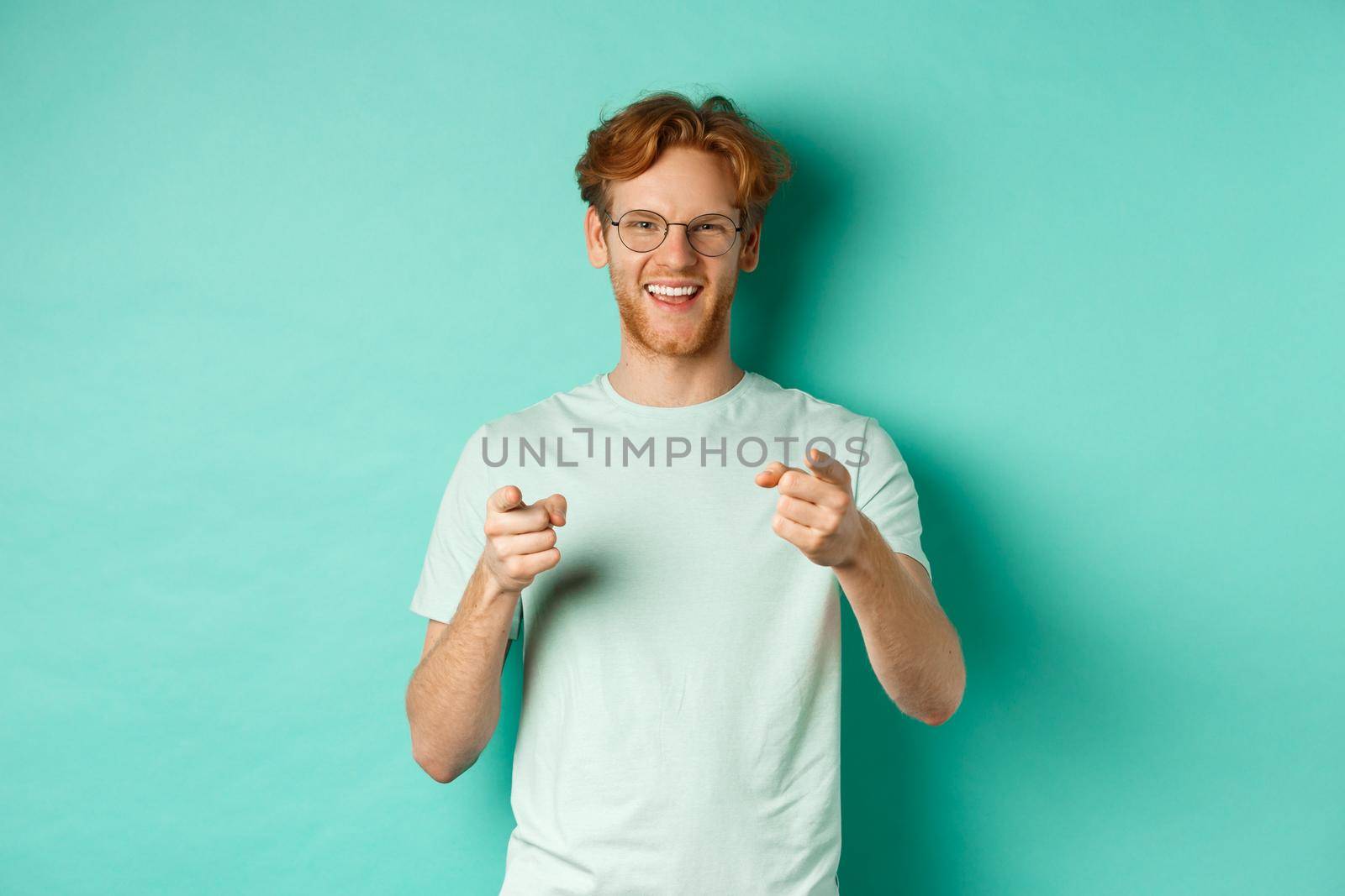 Handsome young man with ginger hair, wearing glasses and t-shirt, pointing finger at camera and smiling, choosing you, congratulating or praising, standing over mint background.