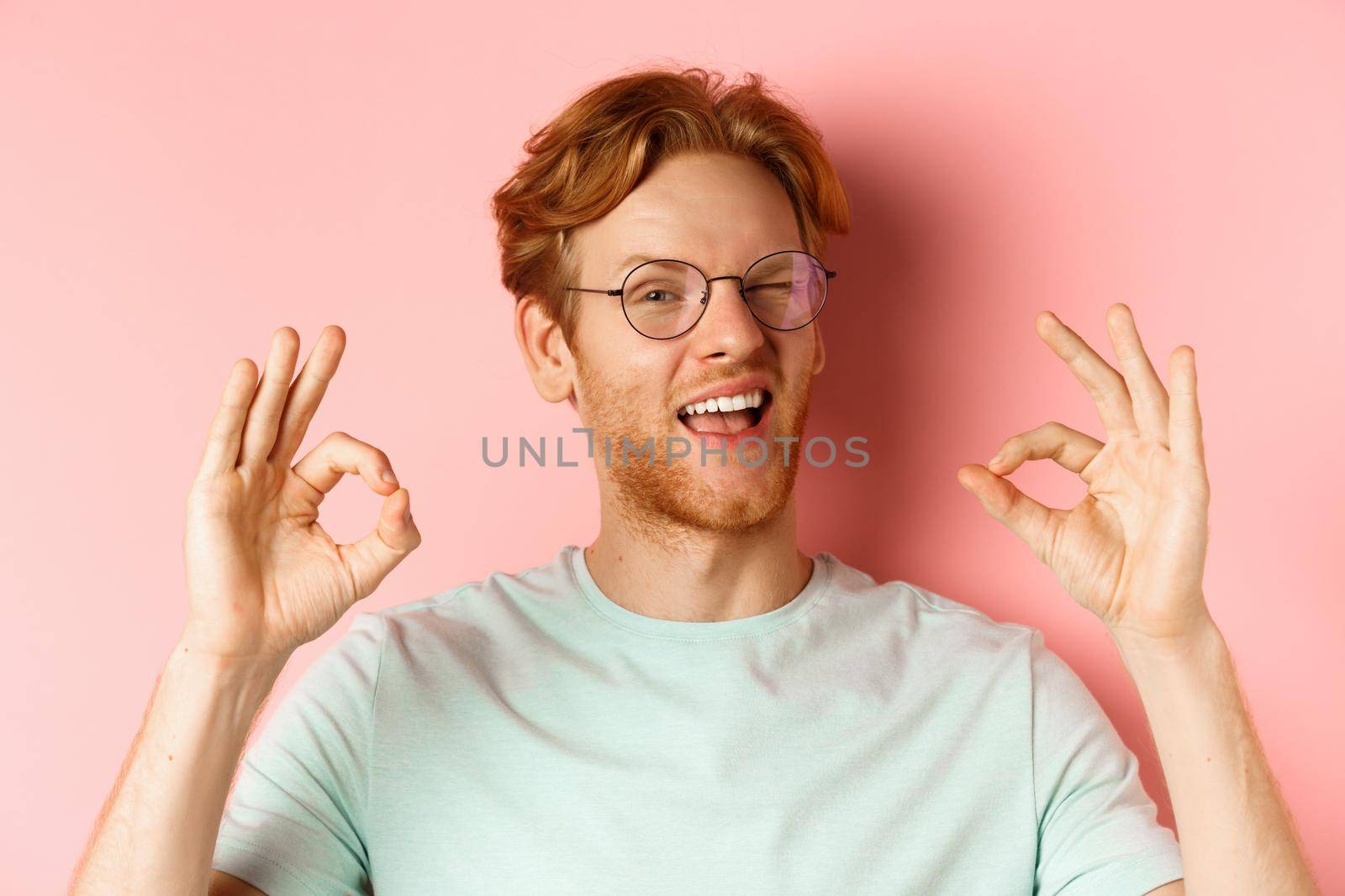 Cheeky redhaed guy in glasses assuring you, winking and showing okay signs, guarantee good quality, standing against pink background.