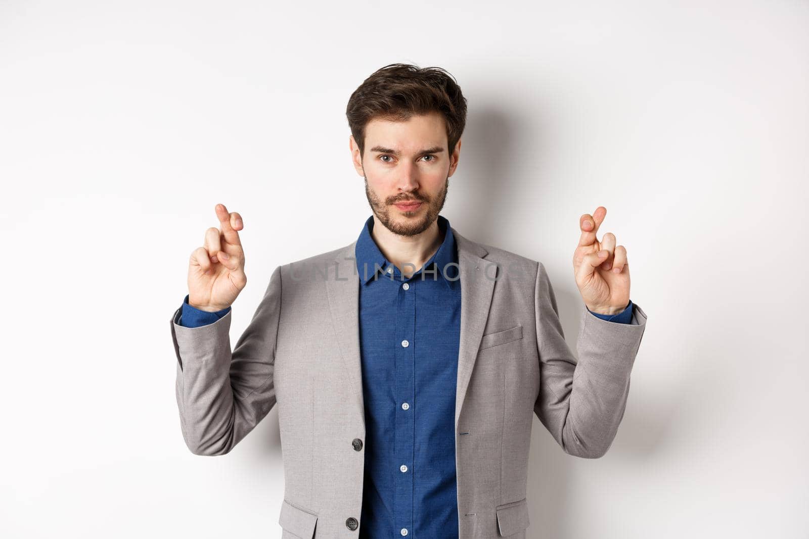 Hopeful man in suit cross fingers for good luck and looking confident in win, feeling determined, standing on white background by Benzoix