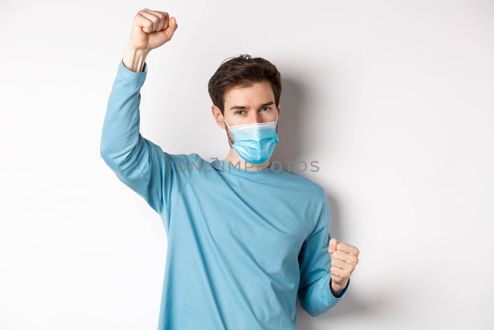 Covid-19, pandemic and social distancing concept. Happy and lucky guy in medical mask winning, raising hands up and celebrating achievement, white background.