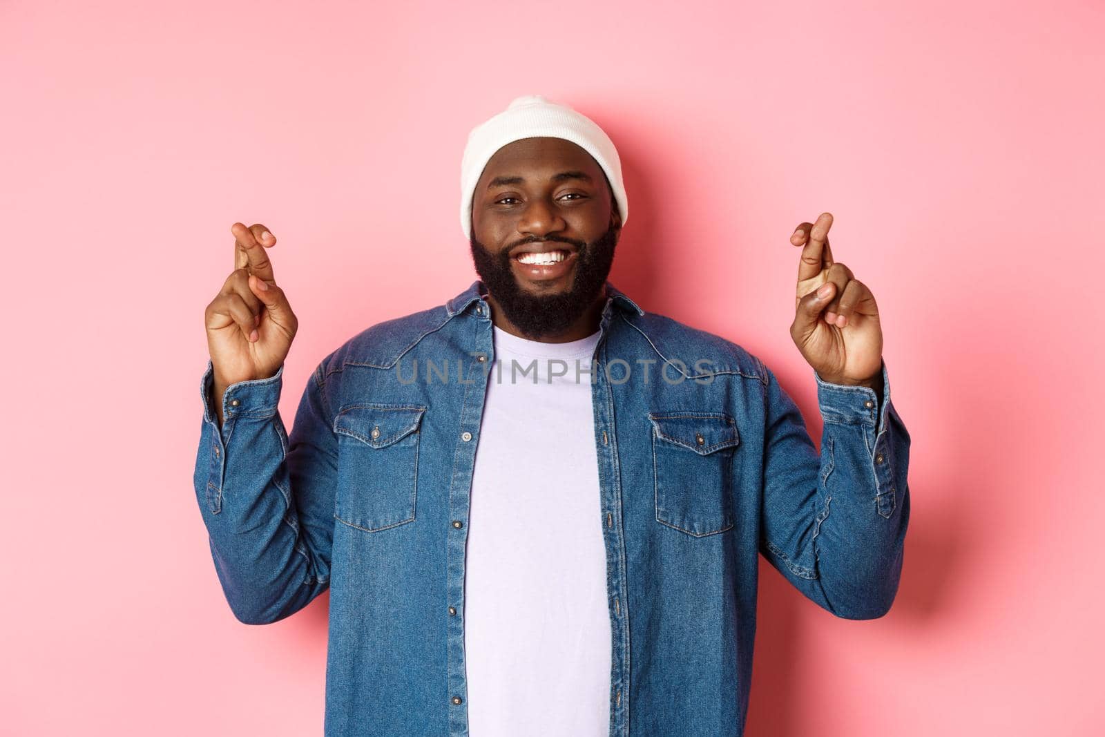 Optimistic african-american man making wish, holding fingers crossed and smiling, standing over pink background.