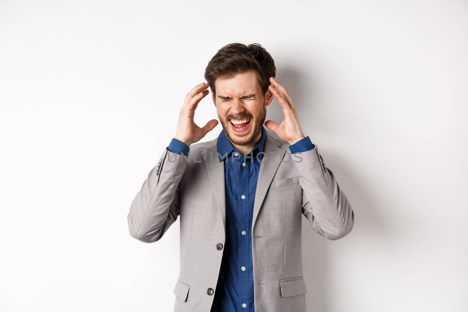 Frustrated business man in suit going crazy, screaming and shaking hands near head, feeling annoyed or angry, shouting distressed, standing on white background.