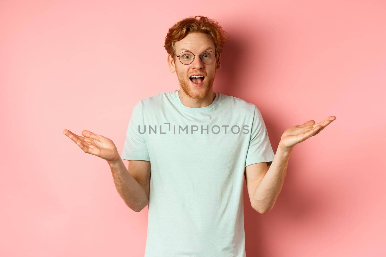 Image of surprised redhead man in glasses, spread hands sideways and looking impressed, standing over pink background.