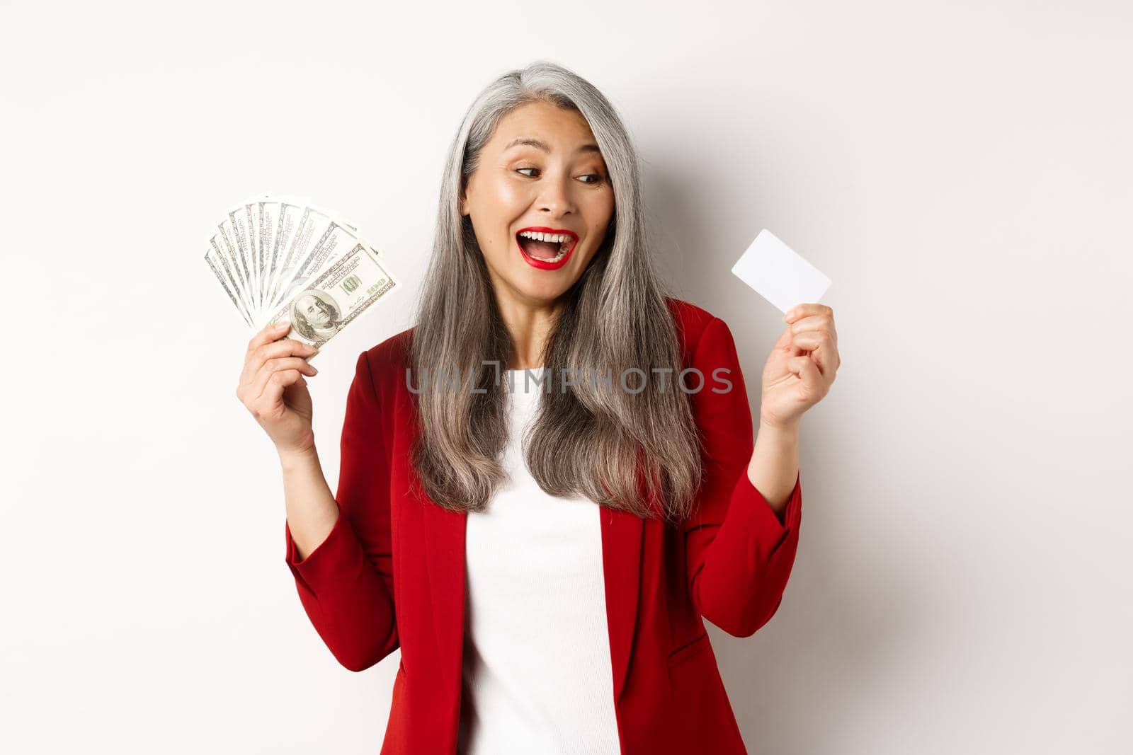 Cheerful asian senior woman earn money, showing dollars in cash and looking at plastic credit card, smiling excited, standing over white background.