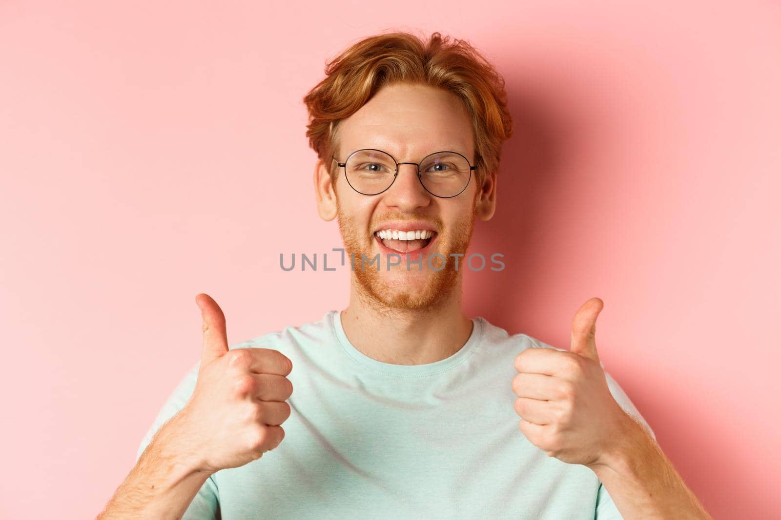 Face of satisfied male customer showing thumbs-up in approval, smiling happy, wearing glasses and t-shirt, pink background.