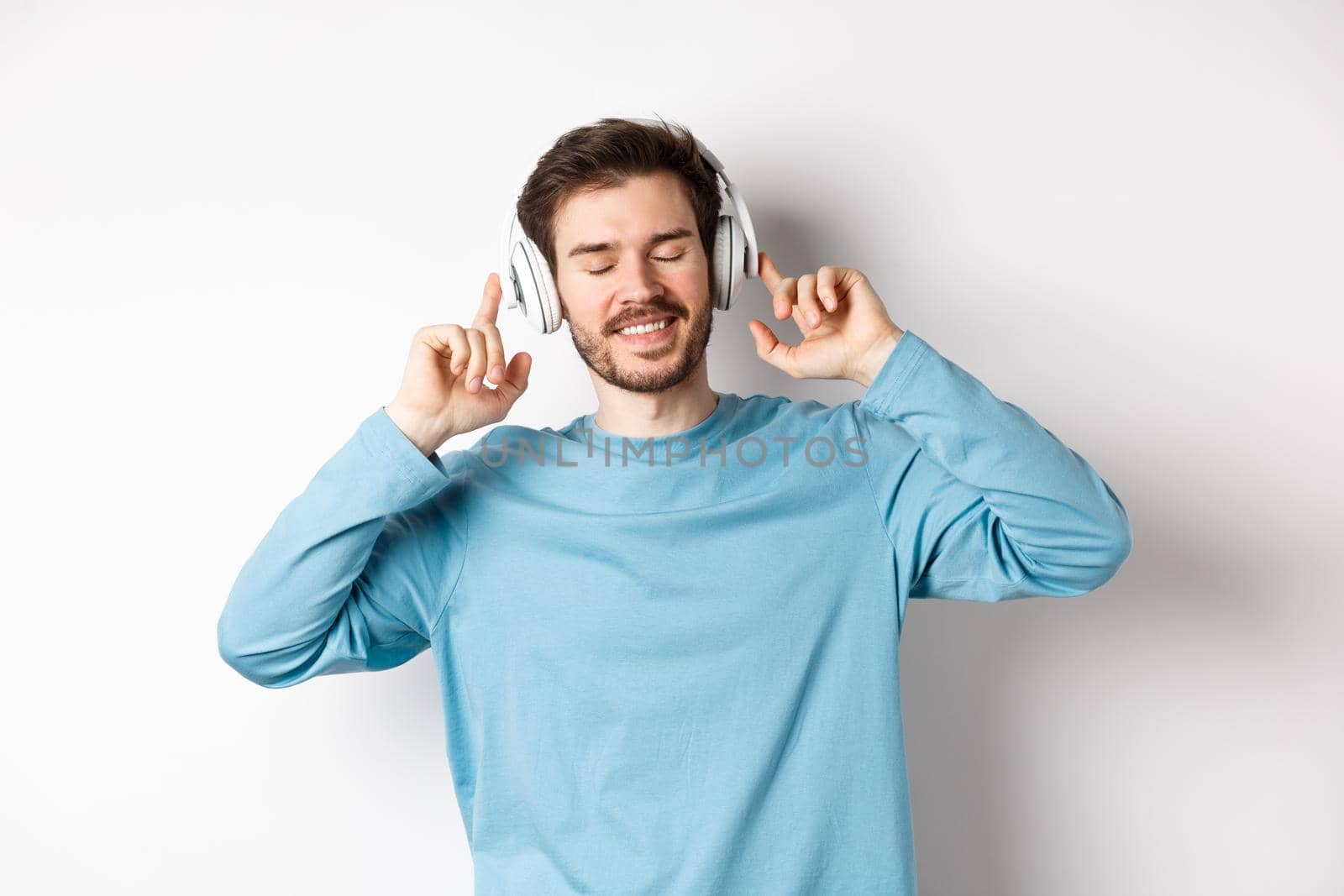 Handsome man listening music in wireless headphones and smiling, enjoying good sound, white background.