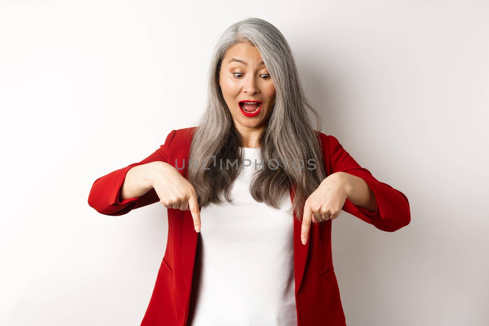 Stylish asian elderly woman in red blazer checking out special promotion, pointing fingers down and looking excited, scream of joy, white background by Benzoix