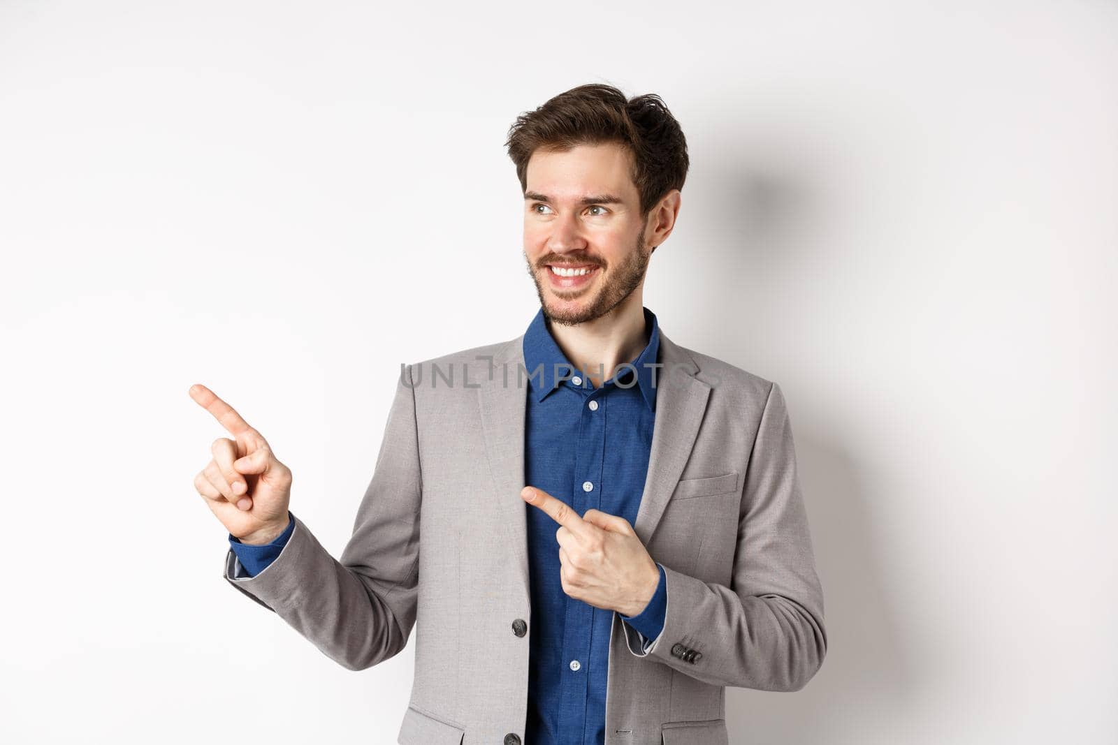 Successful businessman in grey suit pointing fingers left and looking at banner, smiling confident, showing advertisement, standing against white background.