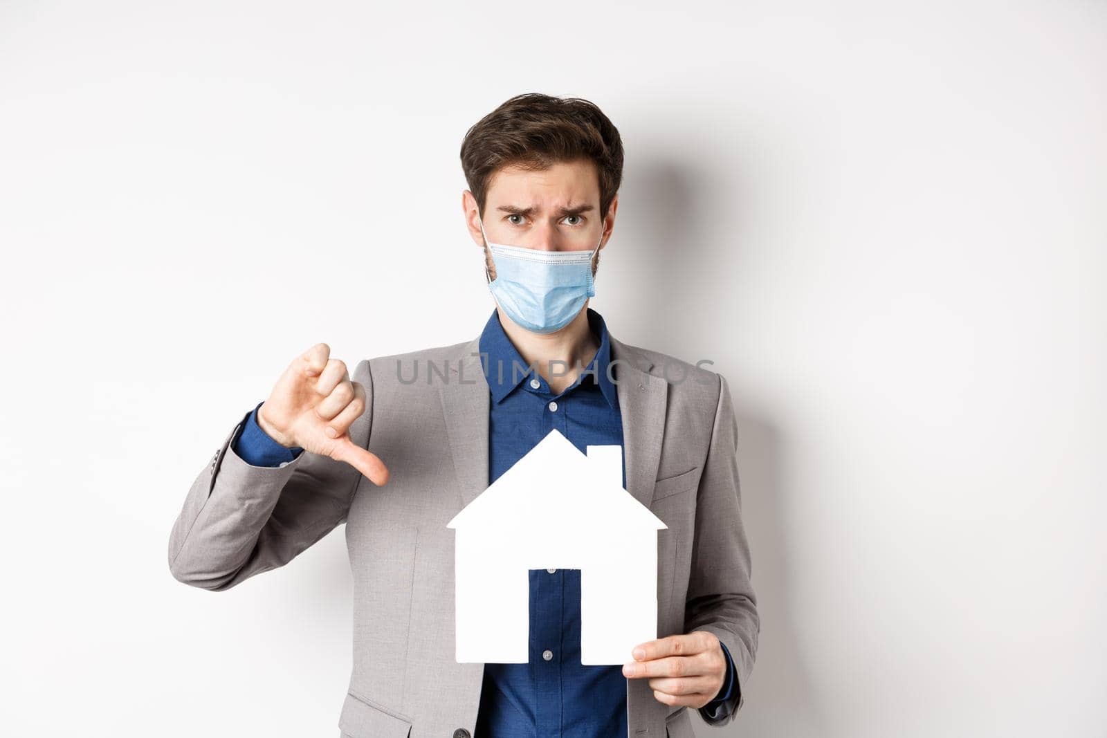 Real estate and covid-19 concept. Disappointed guy in medical mask and suit showing paper house cutout with thumbs-down, complaining on agency, white background.