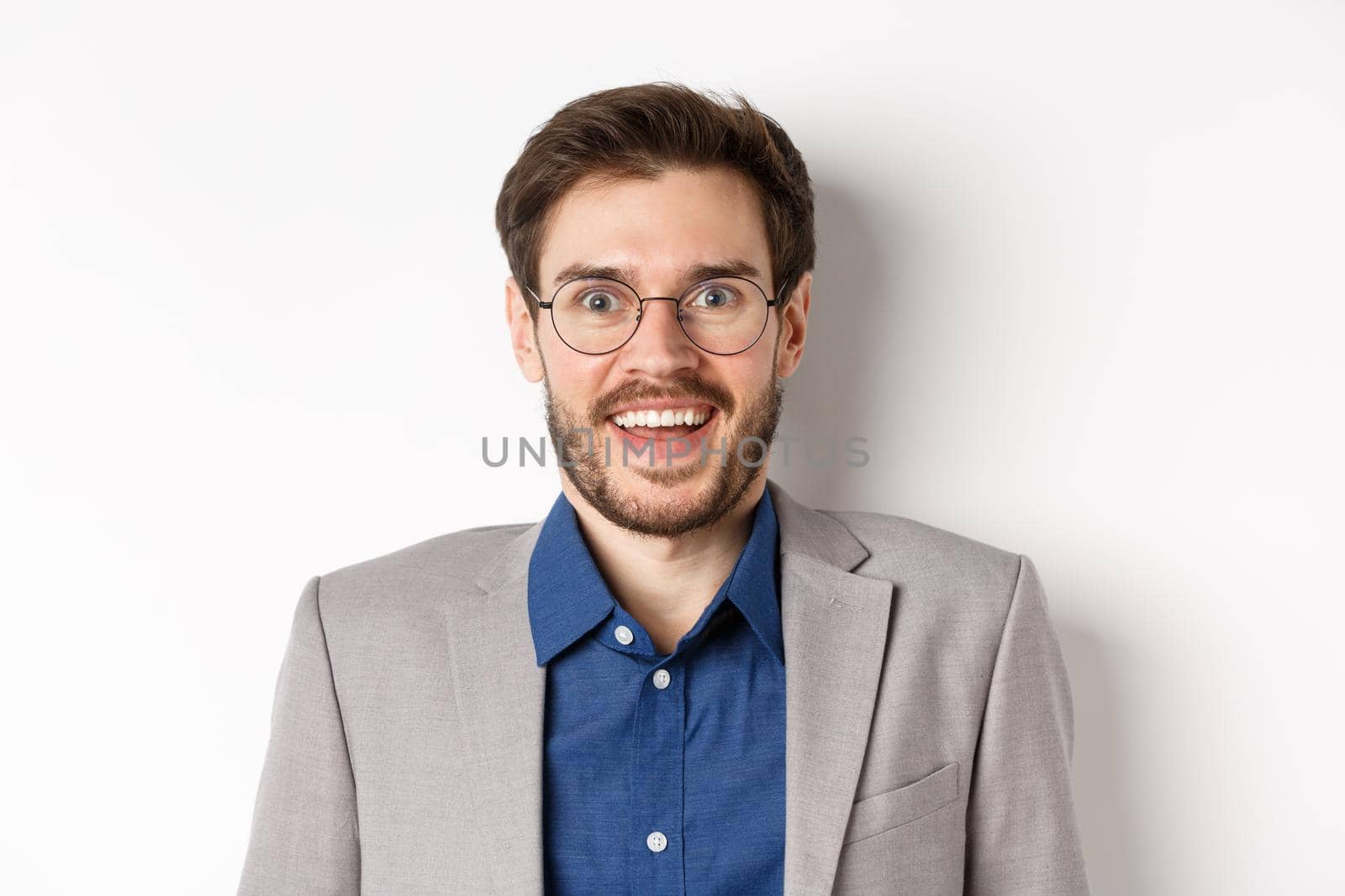 Cheerful european man in glasses and suit smiling excited, looking at camera with amazed happy face, white background.