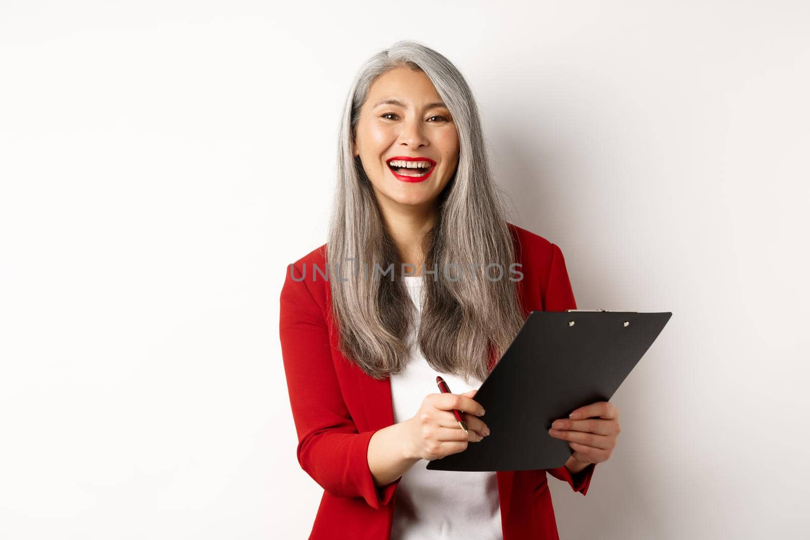 Happy asian office employee, woman in red blazer, working and laughing, holding clipboard with documents, standing over white background.