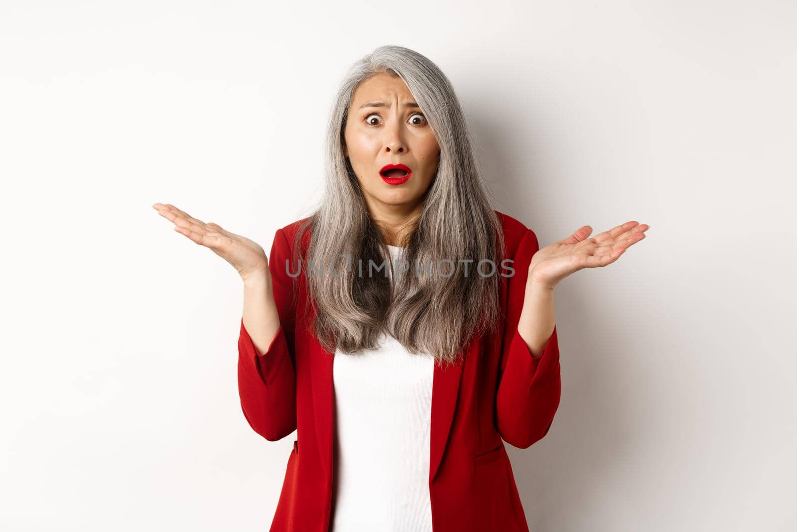 Business people. Confused and anxious asian senior office lady looking worried, spread hands sideways puzzled, standing over white background.
