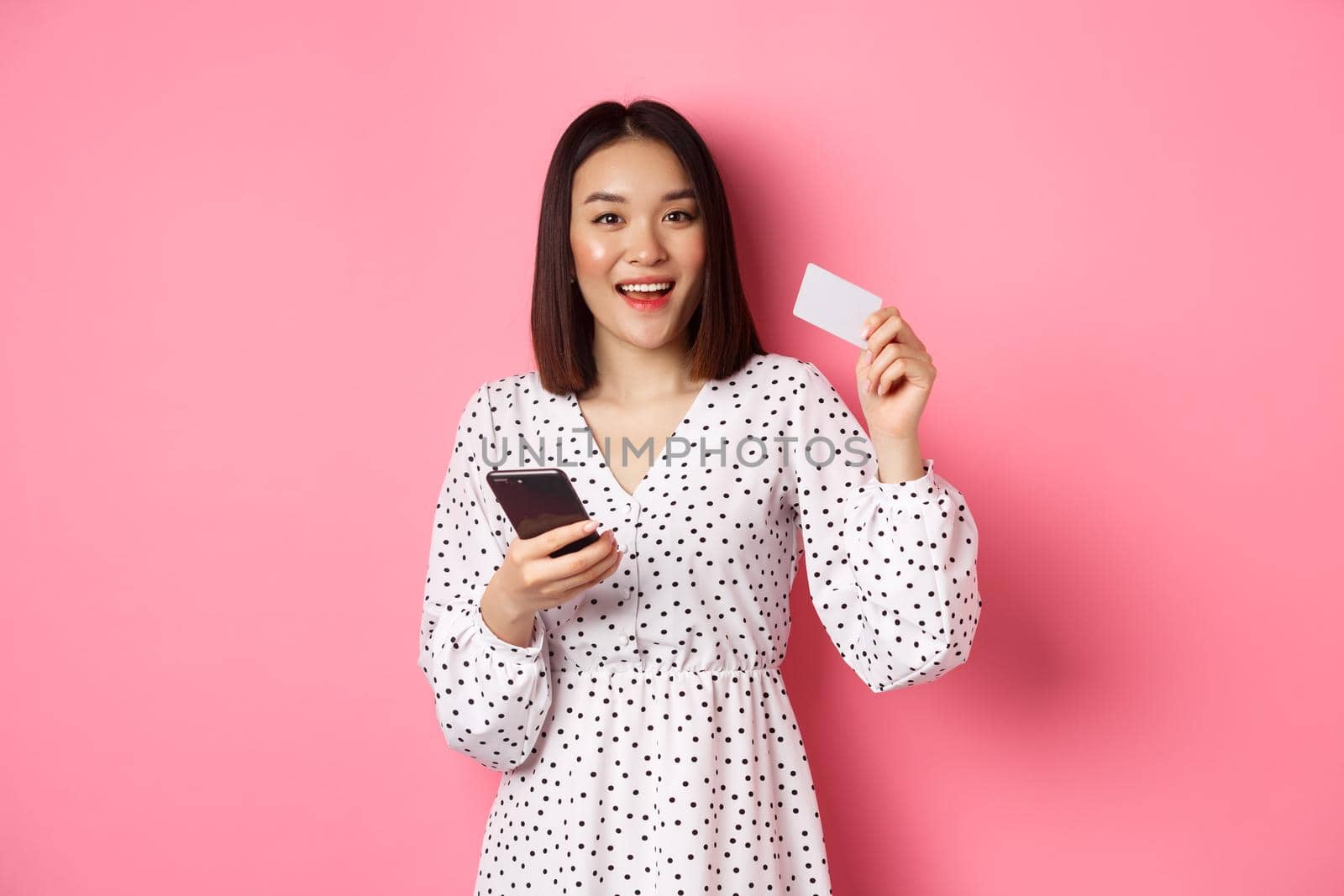 Attractive young asian woman order online, holding credit card and mobile phone, making internet purchase, standing happy over pink background.