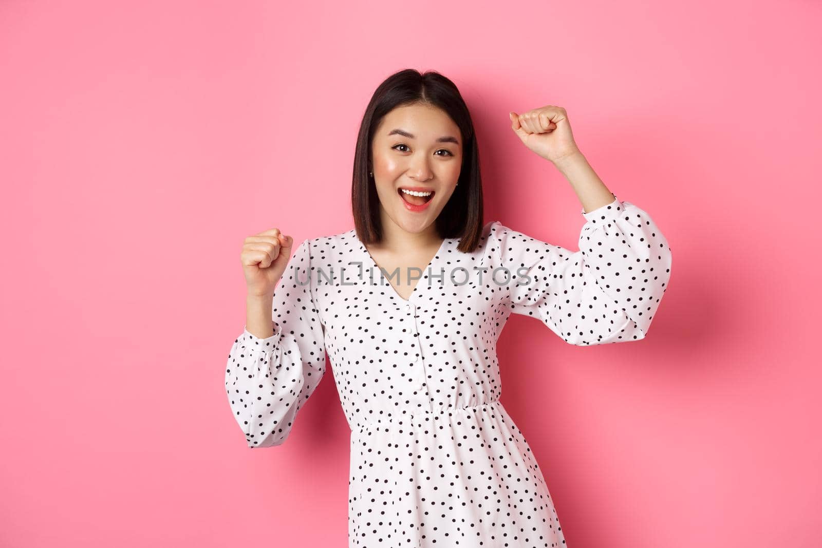 Beautiful korean woman dancing and having fun, smiling happy at camera, posing against pink background.
