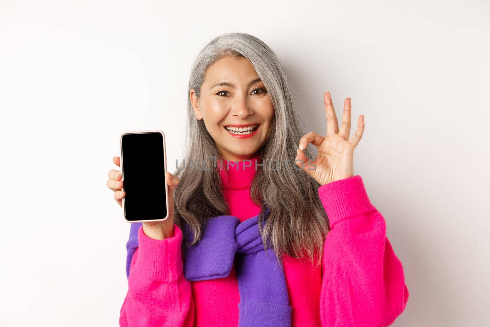 Online shopping. Fashionable asian senior woman showing blank smartphone screen and OK sign, smiling pleased, recommending app, white background.