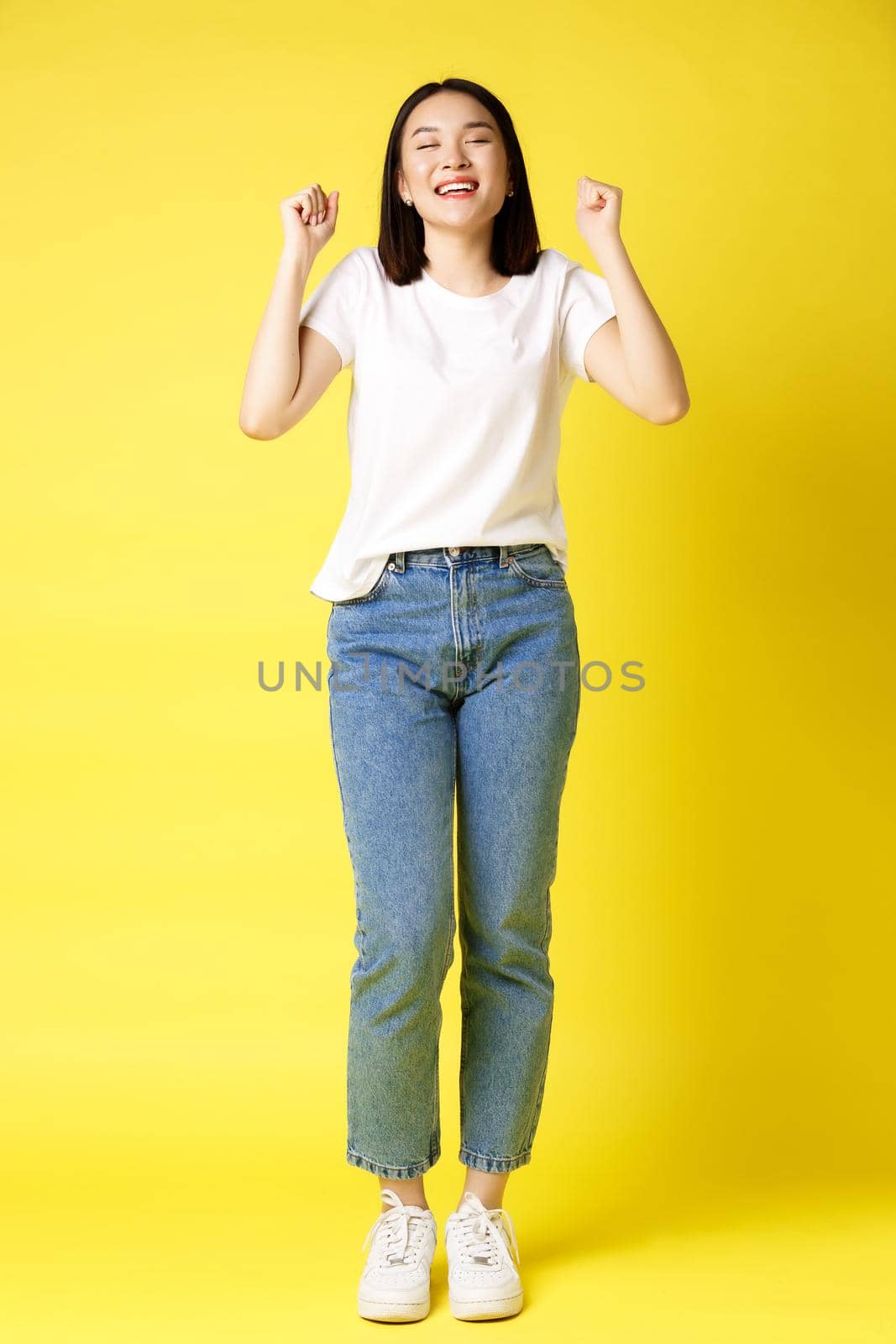 Full size shot of cheerful asian woman in jeans and white t-shirt jumping from happiness, winning prize and rejoicing, celebrating and shouting yes with joy, yellow background.