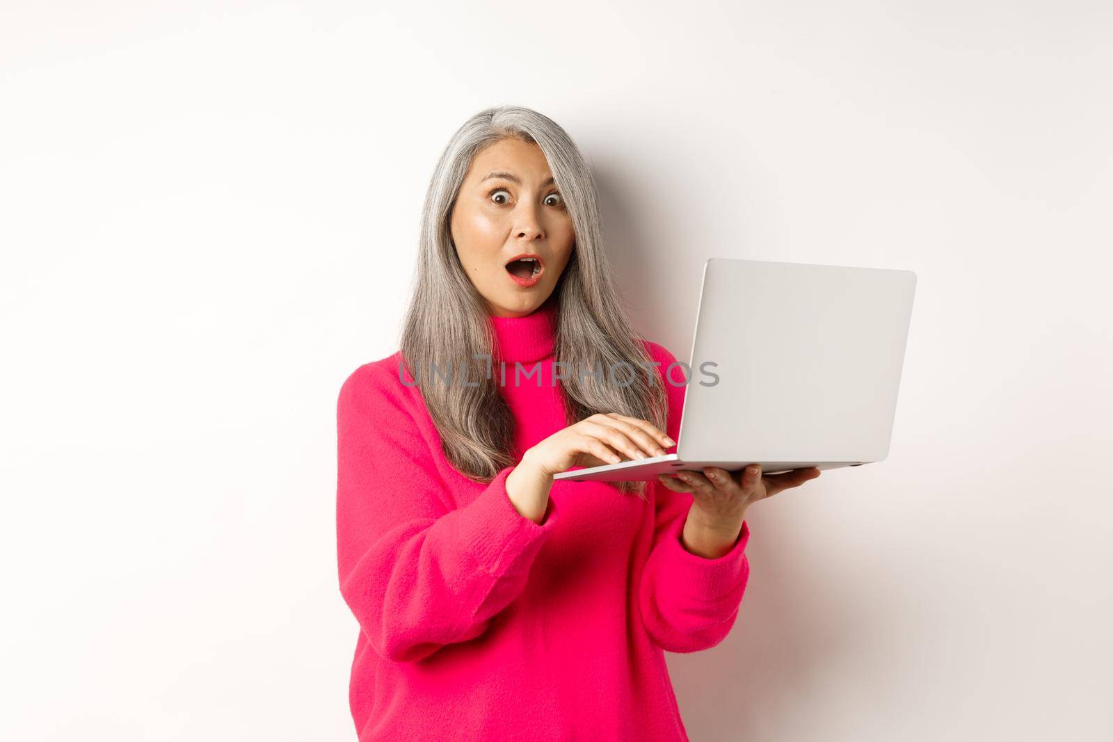 Surprised and impressed asian grandmother staring shocked at camera after reading news on laptop screen, standing over white background.