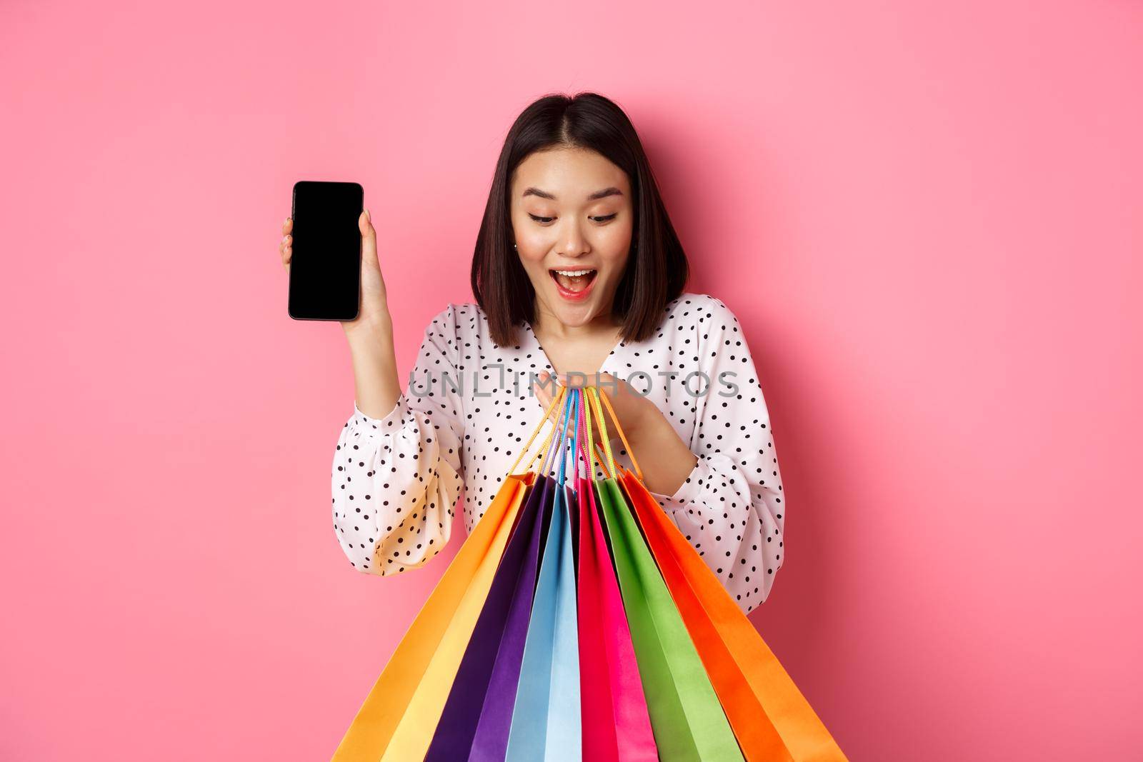 Attractive asian woman showing smartphone app and shopping bags, buying online via application, standing over pink background.