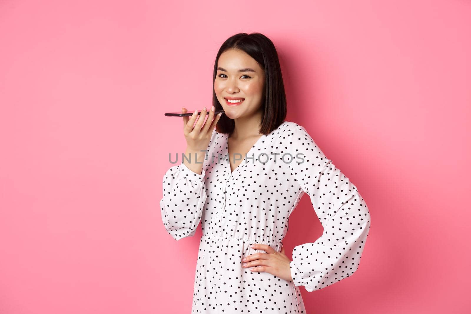 Beautiful korean woman talking on speakerphone, recording voice message and smiling happy, standing over pink background.