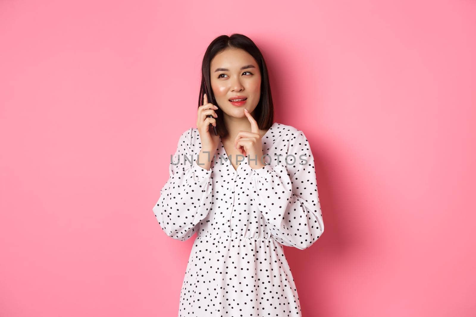 Thoughtful asian woman calling on mobile phone to make order, thinking and looking aside, standing against pink background.