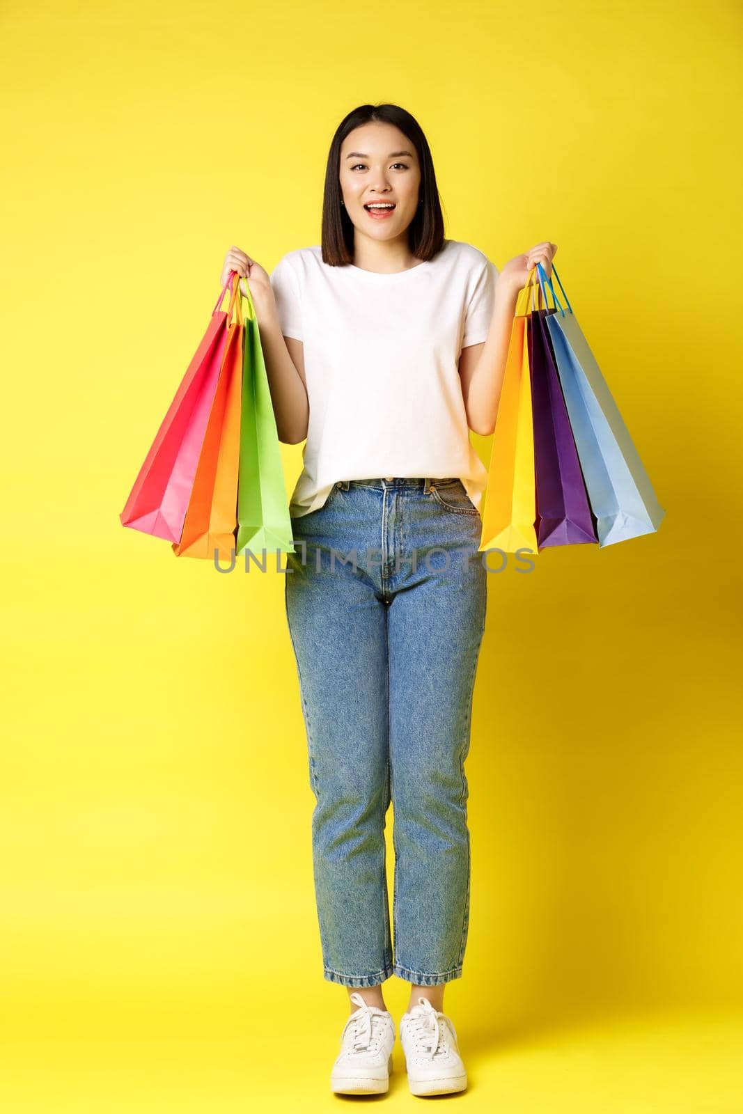 Full size portrait of beautiful asian girl going shopping, holding paper bags from stores and smiling, standing in jeans and white t-shirt over yellow background by Benzoix