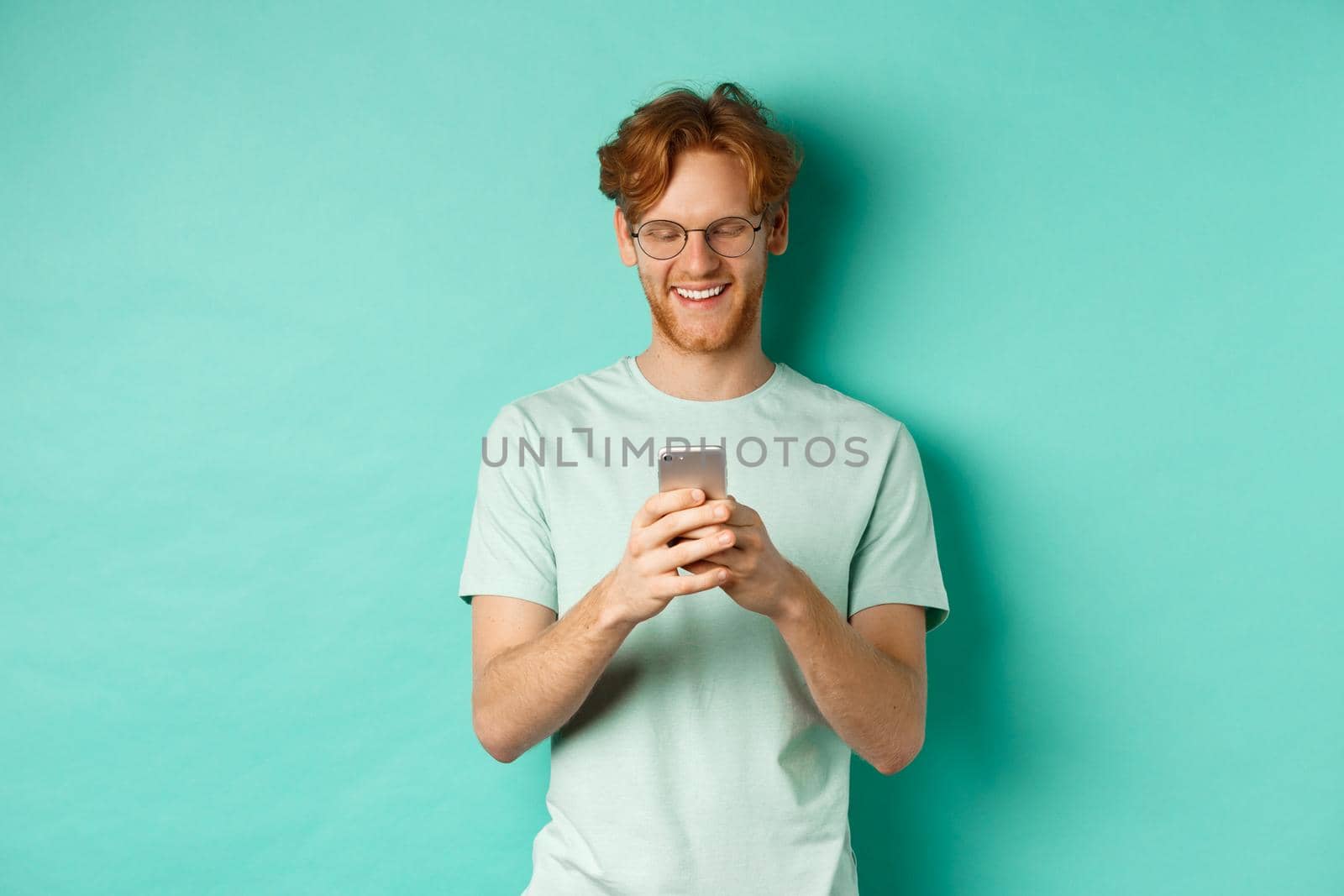 Handsome young man in glasses with red messy hair reading message on mobile phone, smiling and looking at screen, standing over mint background by Benzoix