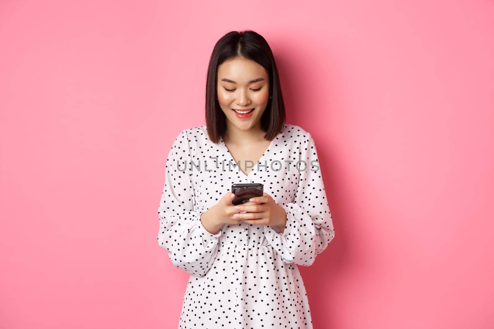 Beautiful asian lady reading message and smiling, using mobile phone, standing in cute dress against pink background.