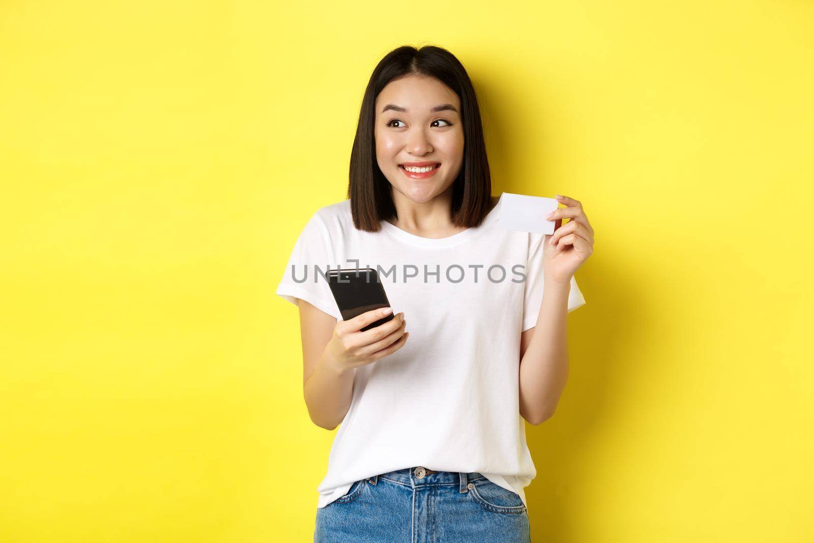 E-commerce and online shopping concept. Cheerful asian girl paying in internet, holding smartphone and plastic credit card, smiling and looking left, yellow background.