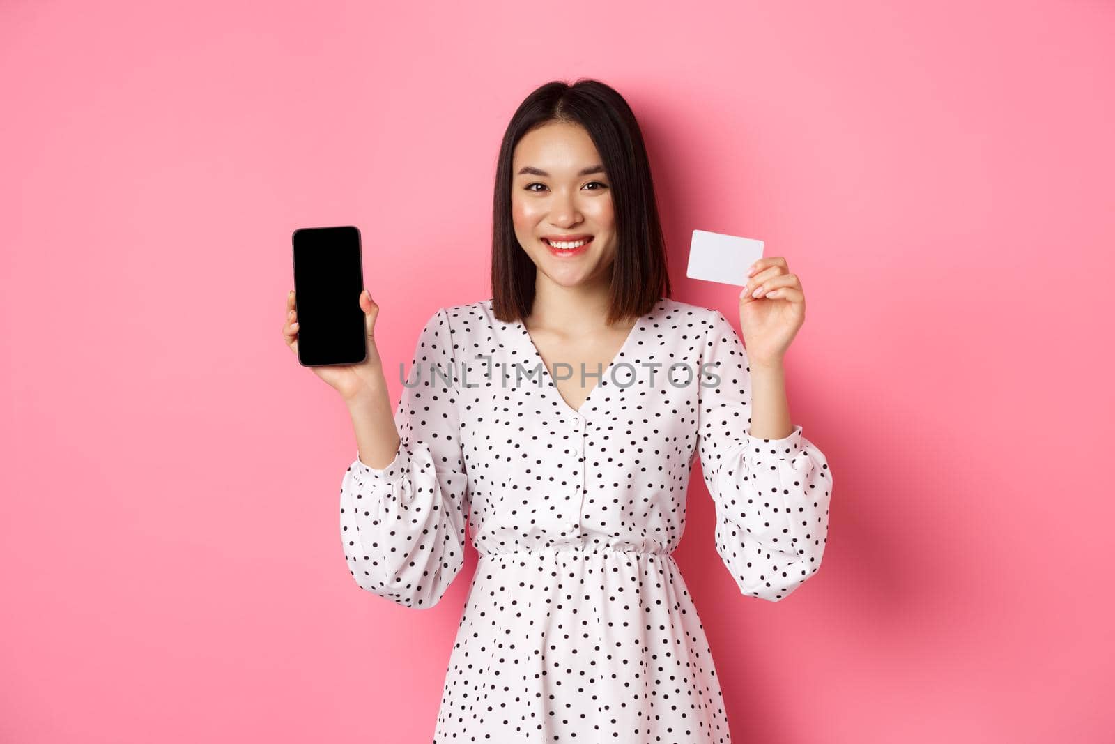 Cute asian woman shopping online, showing bank credit card and mobile screen, smiling and looking at camera, standing over pink background.