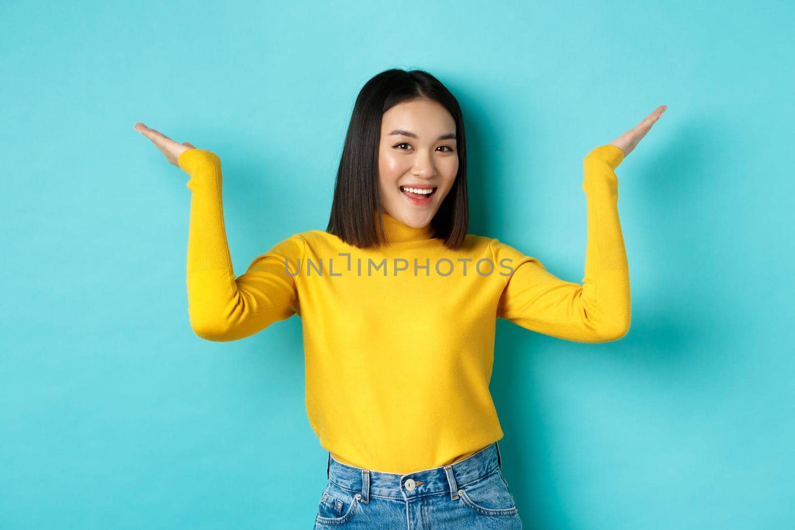 Beauty and fashion concept. Stylish beautiful asian girl demonstrate promotion, raising hands up and smiling, showing something on blue background.