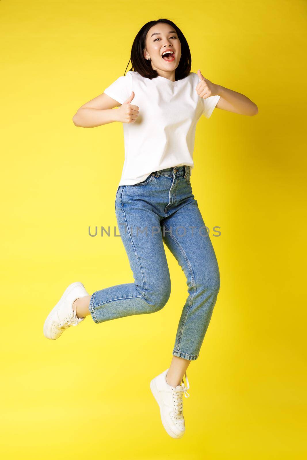 Full size of happy young asian woman jumping from joy, showing thumbs up in approval, posing over yellow background in jeans and casual white t-shirt by Benzoix