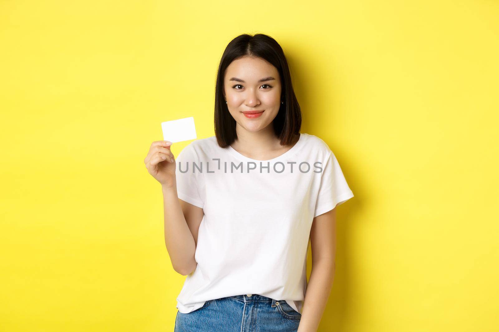 Young asian woman in casual white t-shirt showing plastic credit card and smiling at camera, yellow background by Benzoix