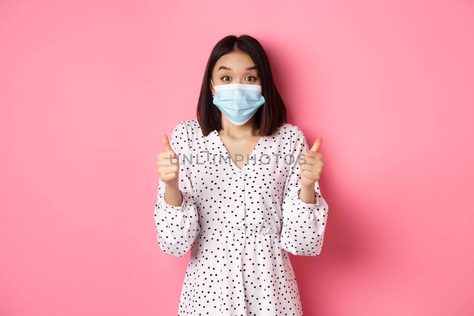 Covid-19, quarantine and lifestyle concept. Excited asian woman in face mask showing thumbs-up, praising good job, looking amazed at camera, standing over pink background by Benzoix