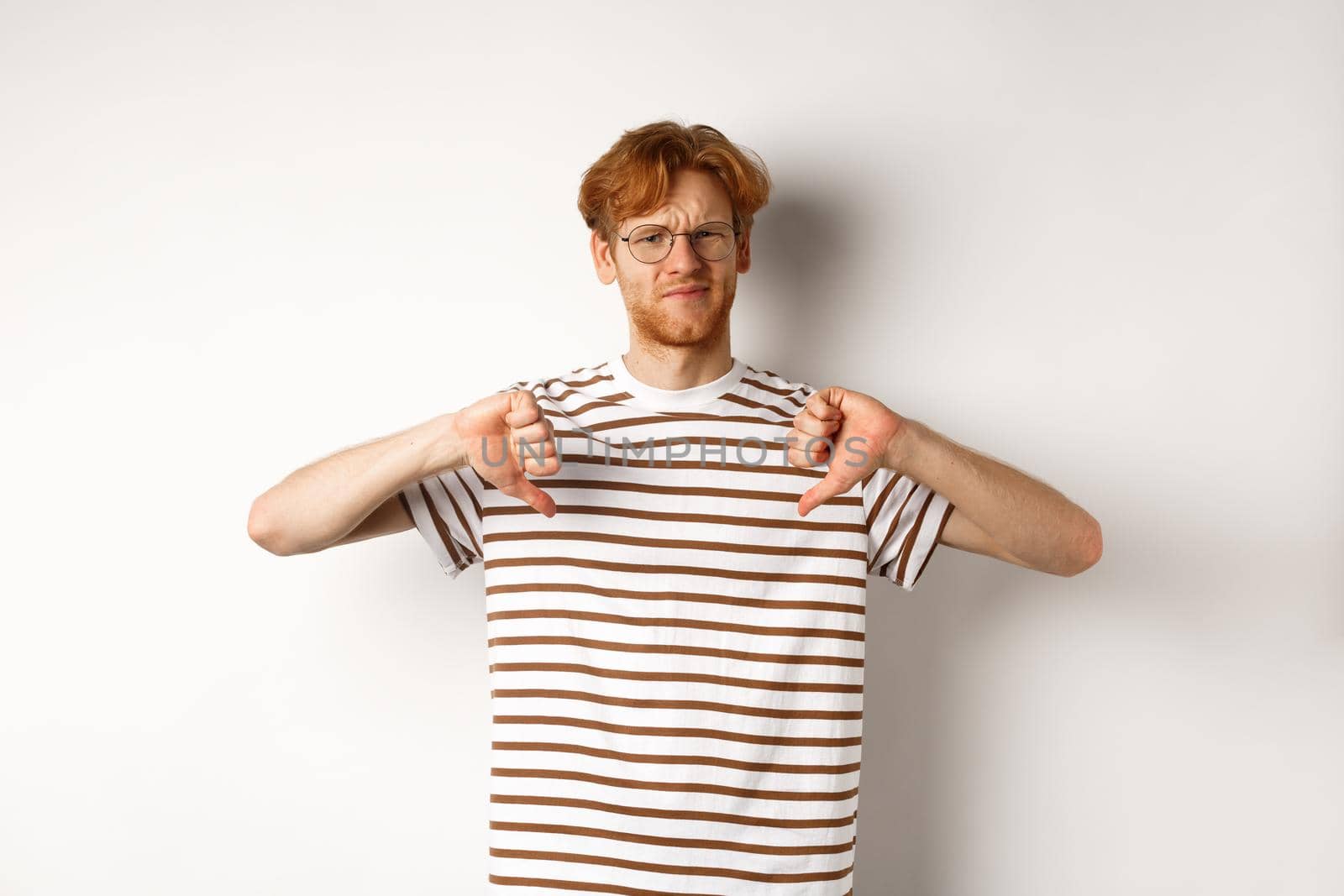 Disappointed young man with red hair and glasses disagree, showing thumbs-down and frowning displeased, standing over white background.