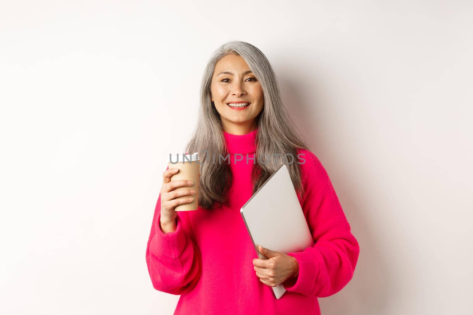 E-commerce concept. Happy asian senior freelancer, drinking coffee and holding laptop, smiling relaxed at camera, standing over white background.