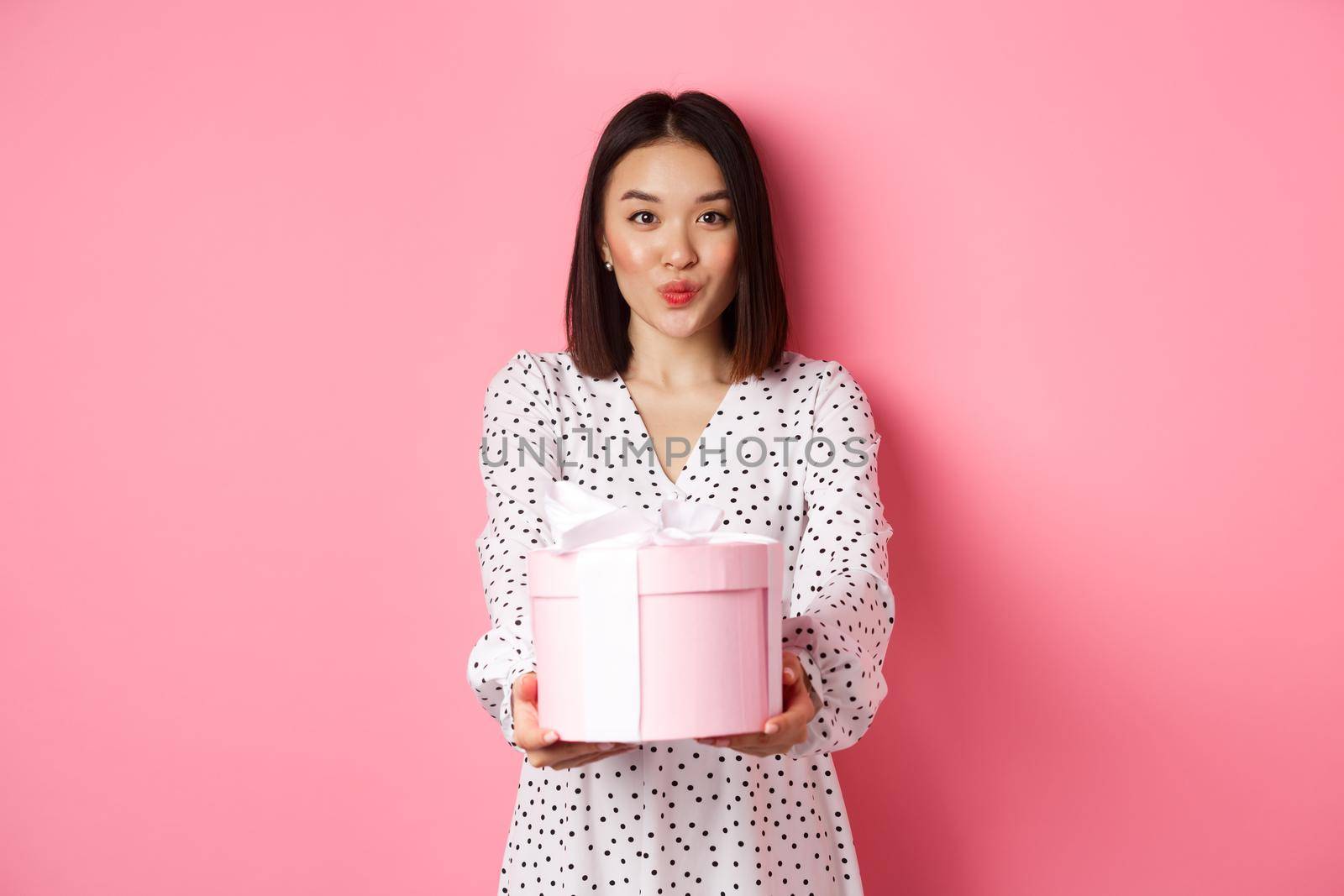 Cute asian girlfriend congratulate with valentines day, giving cute romantic gift in box, pucker lips for kiss, standing over pink background.