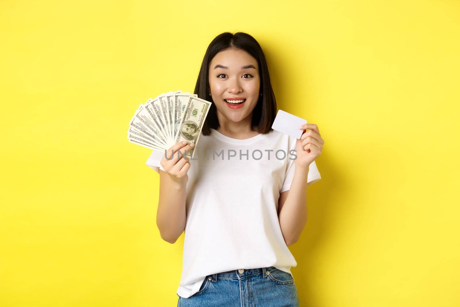Beautiful asian woman with short dark hair, wearing white t-shirt, showing money in dollars and plastic credit card, standing over yellow background.