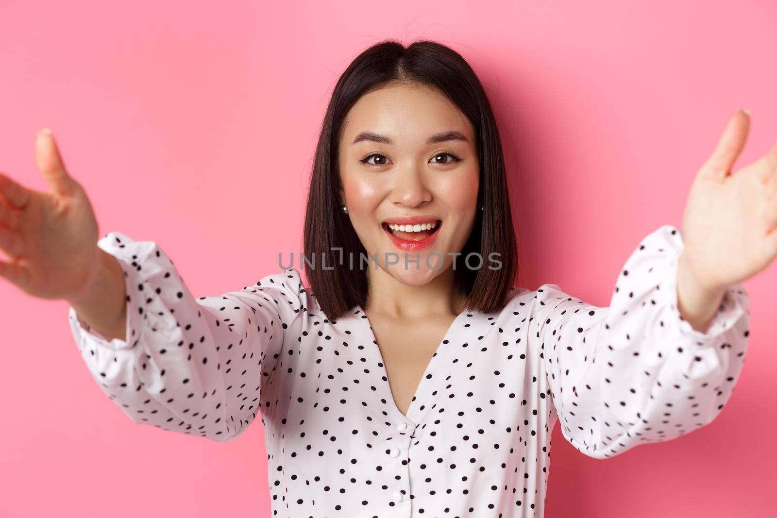 Close-up of cute asian woman reaching hands forward, taking selfie, record beauty blog and smiling, standing over pink background.