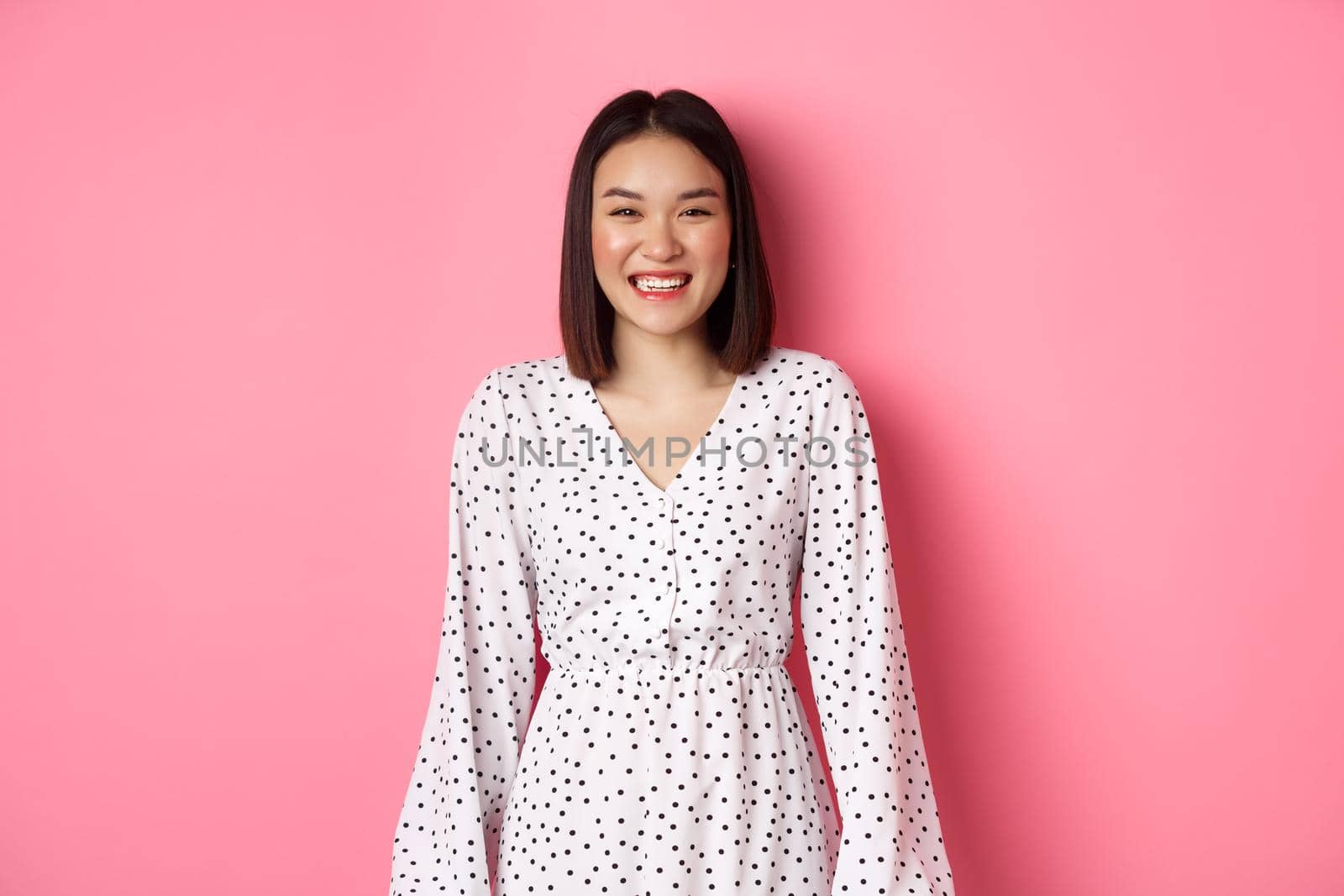 Happy korean woman in dress looking at camera, smiling and laughing with sincere expression, standing over pink background. Copy space