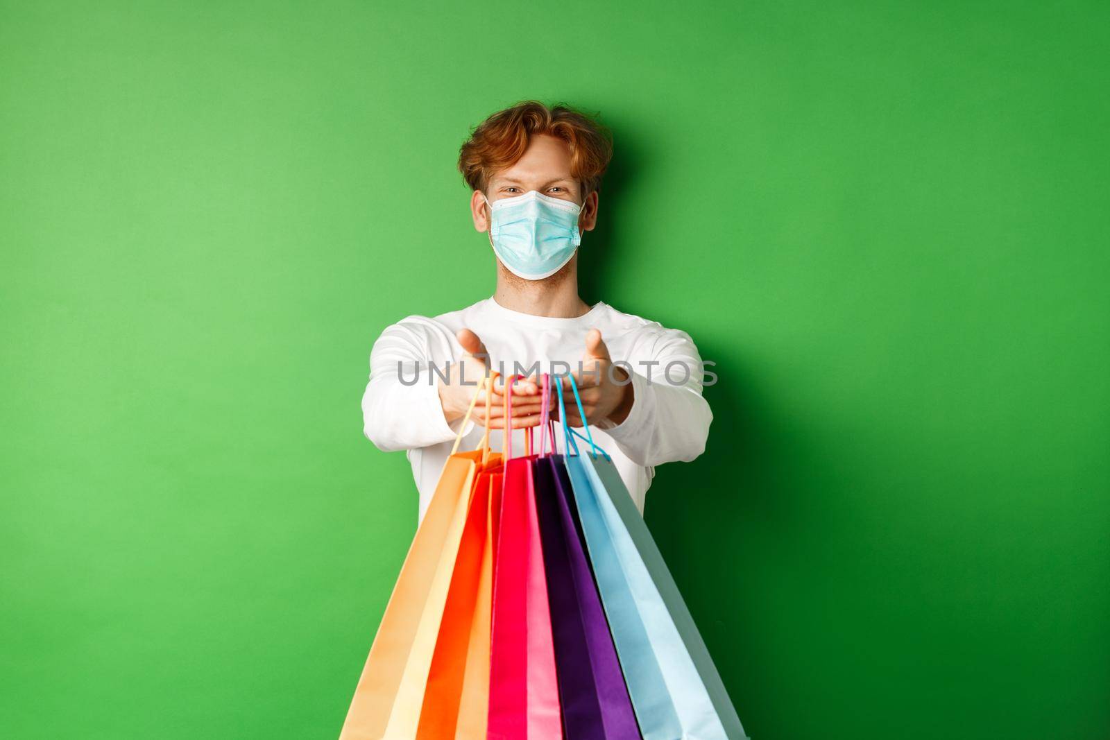 Happy young man in medical mask giving you shopping bags with purchases, smiling and wishing well, standing over green background. Covid-19 concept.